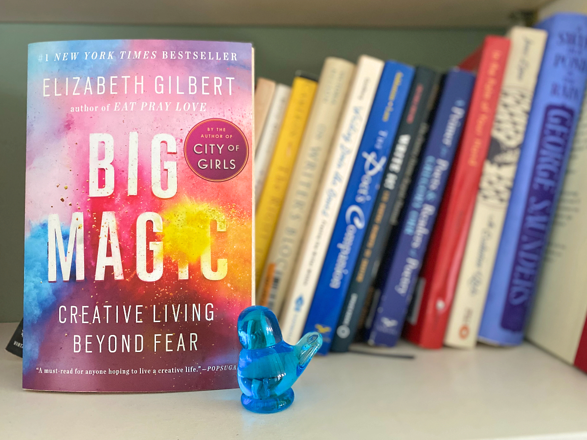 The book Big Magic by Elizabeth Gilbert sits face-forward on a shelf with several books about writing poetry and a small, blue glass statue of a bird.