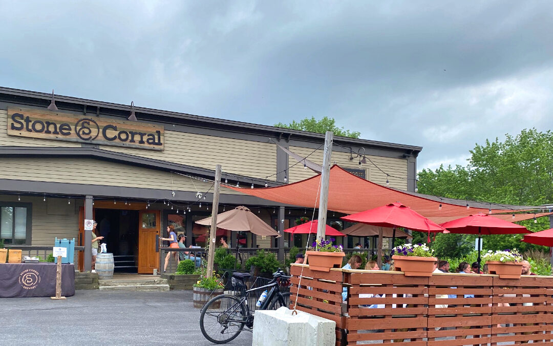 A one-story building with light brown paneling and sign that says Stone Corral in an old West-style font. In front are picnic tables with red umbrellas.