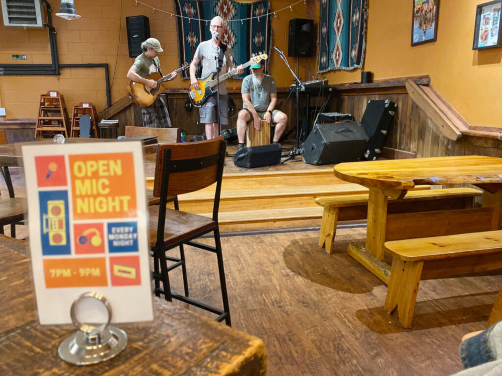 Three men with gray hair play instruments on stage in the back corner of a restaurant. There are wooden picnic tables in the foreground and a table tent that says Open Mic Night Every Monday Night 7-9pm.